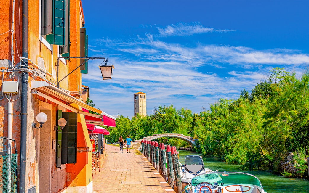 Visitors walking along a canal in Torcello Island with a view of a historic tower and bridge.
