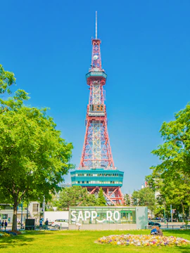 Sapporo TV Tower in Odori Park, surrounded by trees and flowers, under a clear blue sky.