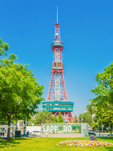 Sapporo TV Tower in Odori Park, surrounded by trees and flowers, under a clear blue sky.