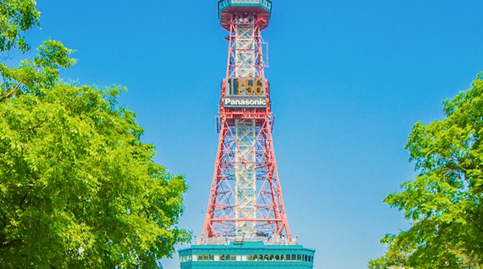 Sapporo TV Tower in Odori Park, surrounded by trees and flowers, under a clear blue sky.