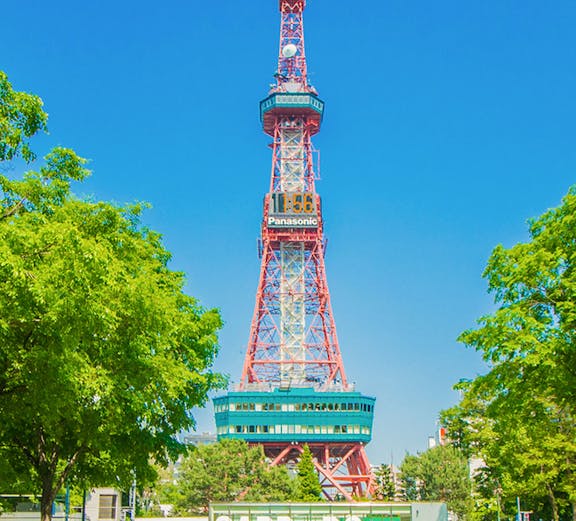 Sapporo TV Tower in Odori Park, surrounded by trees and flowers, under a clear blue sky.