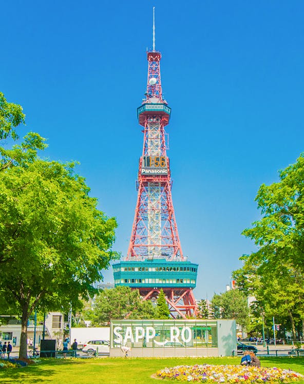 Sapporo TV Tower in Odori Park, surrounded by trees and flowers, under a clear blue sky.