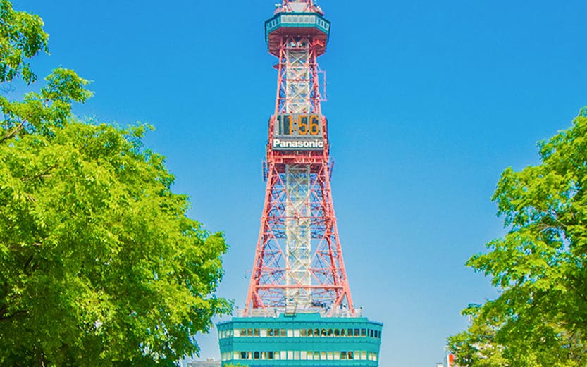 Sapporo TV Tower in Odori Park, surrounded by trees and flowers, under a clear blue sky.