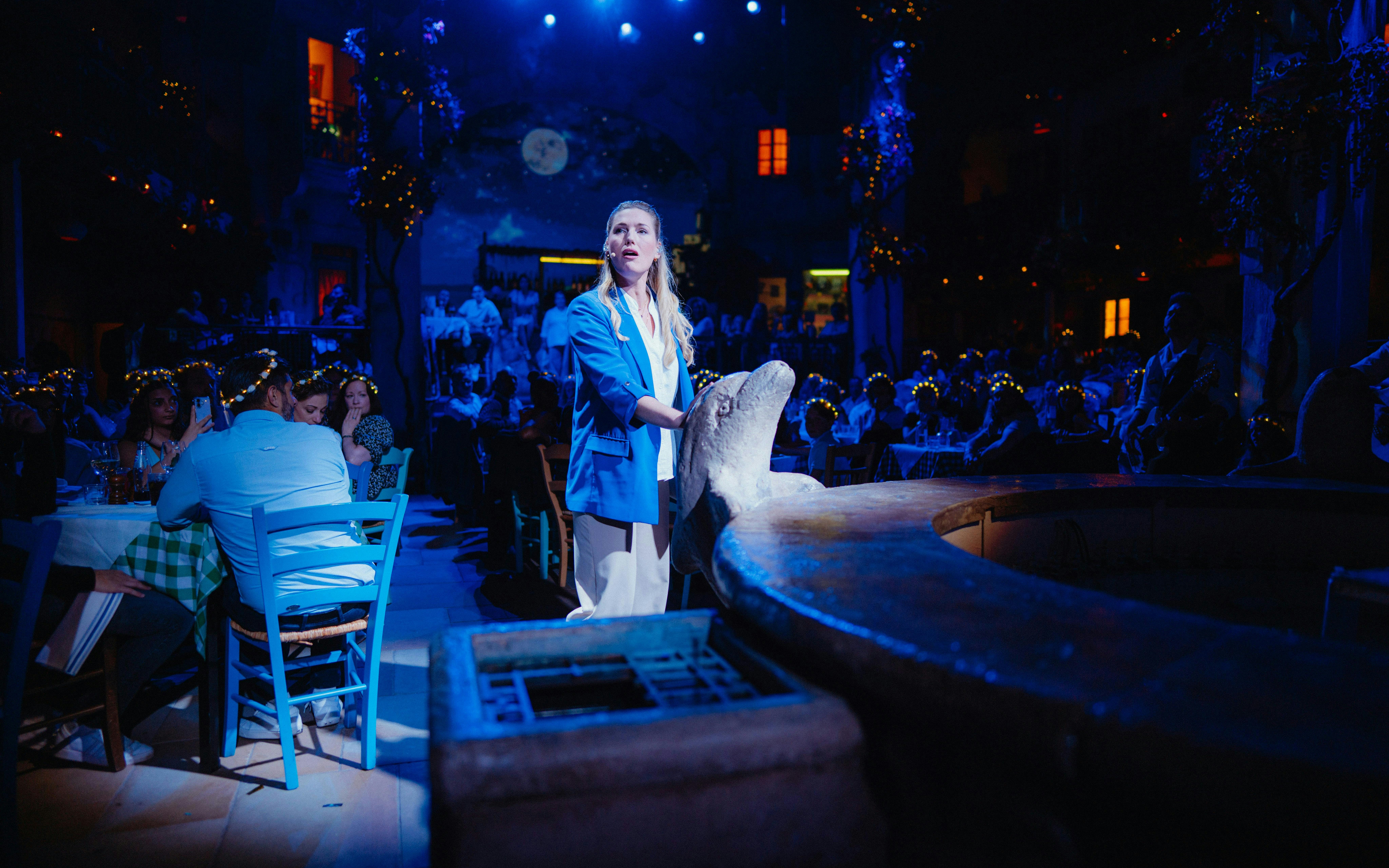 Performer singing during Mamma Mia! The Party with audience seated at tables.