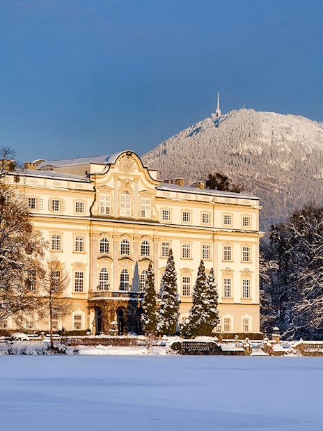 Leopoldskron Palace in winter with snow-covered trees and mountain backdrop.