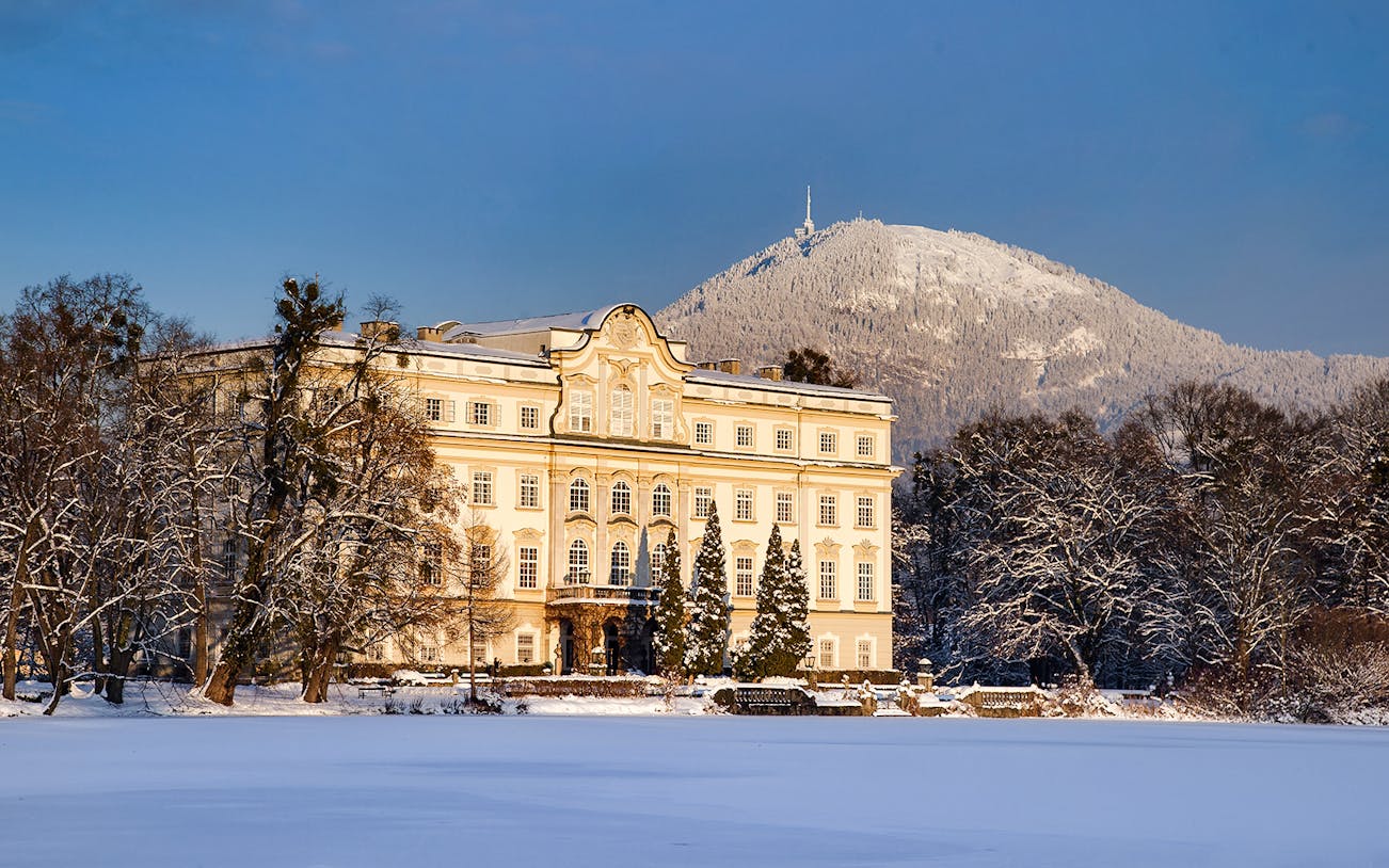 Leopoldskron Palace in winter with snow-covered trees and mountain backdrop.