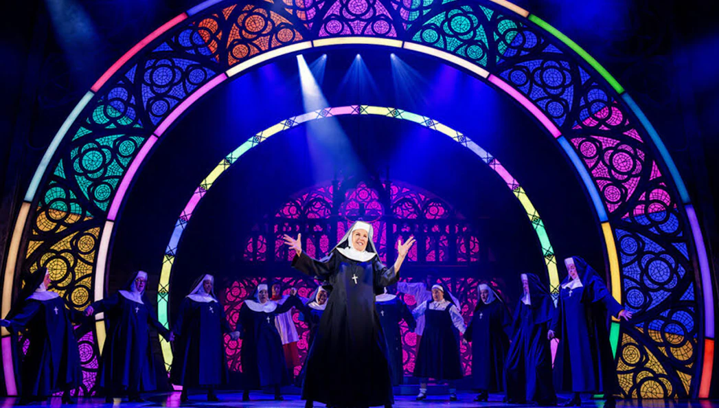 Nuns performing on stage in a scene from the musical "Sister Act" with colorful stained glass backdrop.