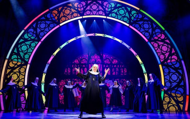 Nuns performing on stage in a scene from the musical "Sister Act" with colorful stained glass backdrop.
