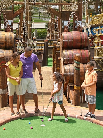 Family playing mini-golf at Pirate's Cove Adventure Golf with pirate ship backdrop.