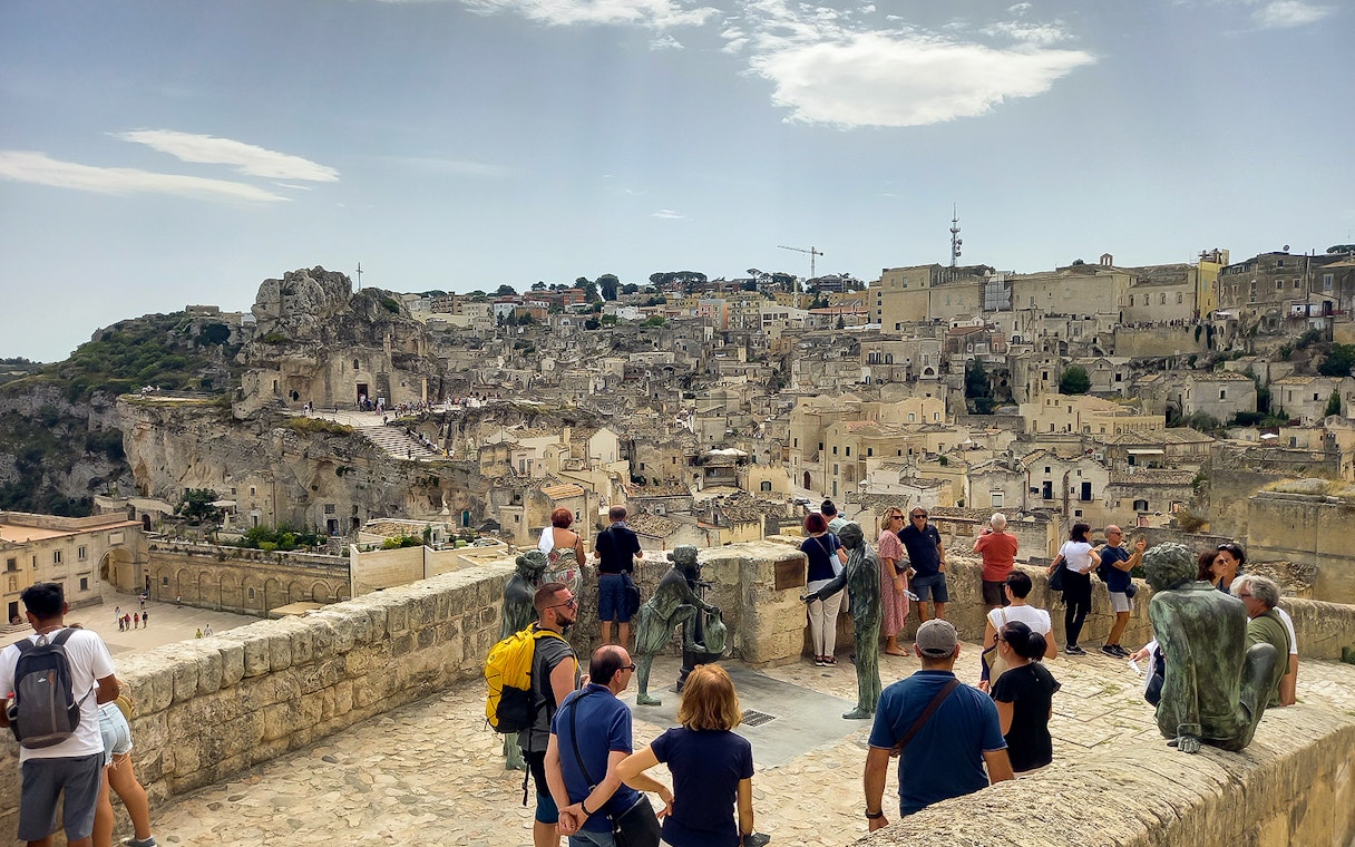 Tourists overlooking Matera's ancient cityscape with stone buildings and sculptures.