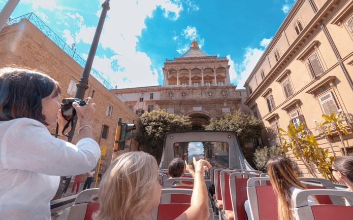 Sightseers on an open-top bus in Palermo photographing historic architecture.