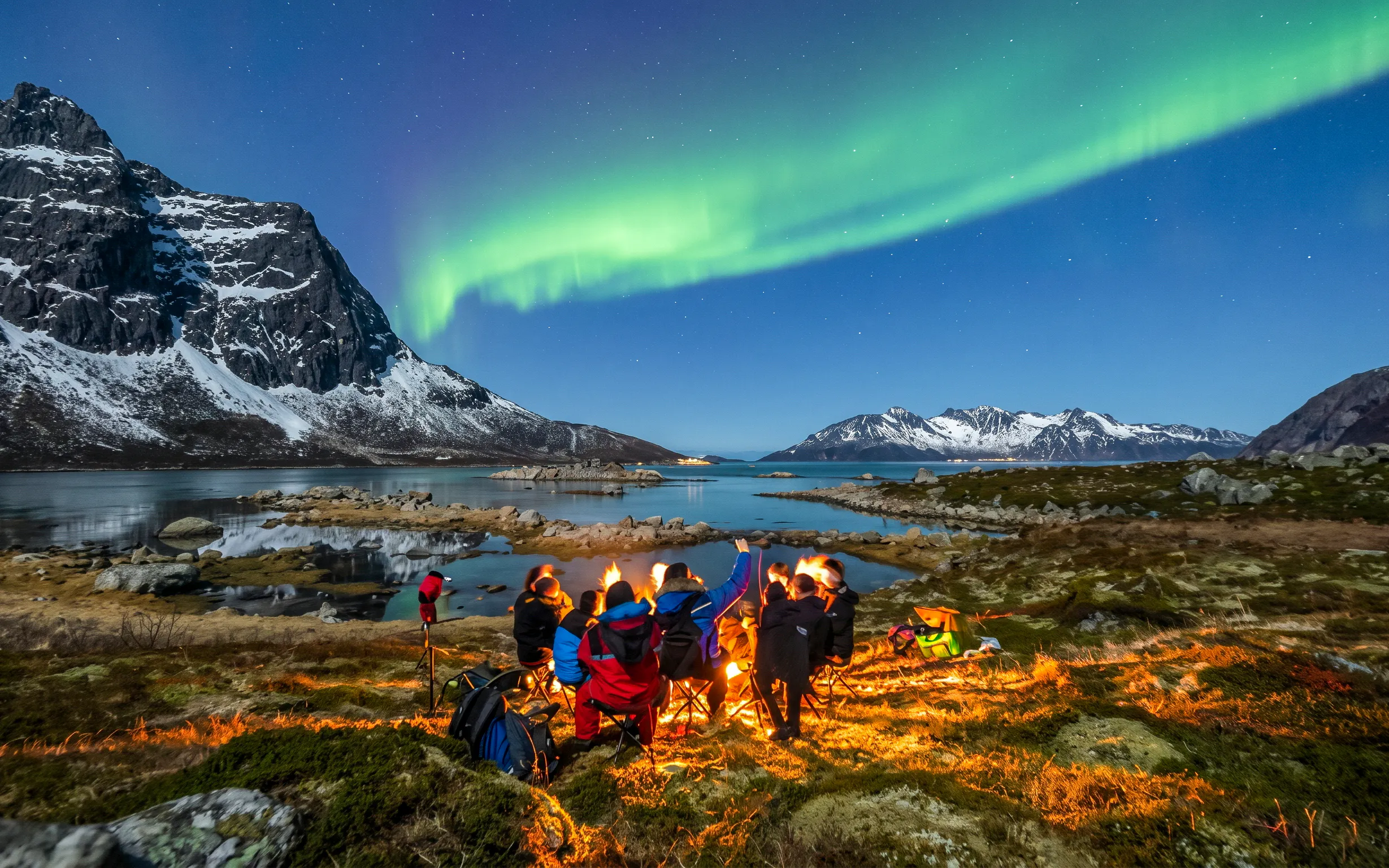 Group watching Northern Lights by a campfire in Tromsø, Norway, with mountains and water in the background.