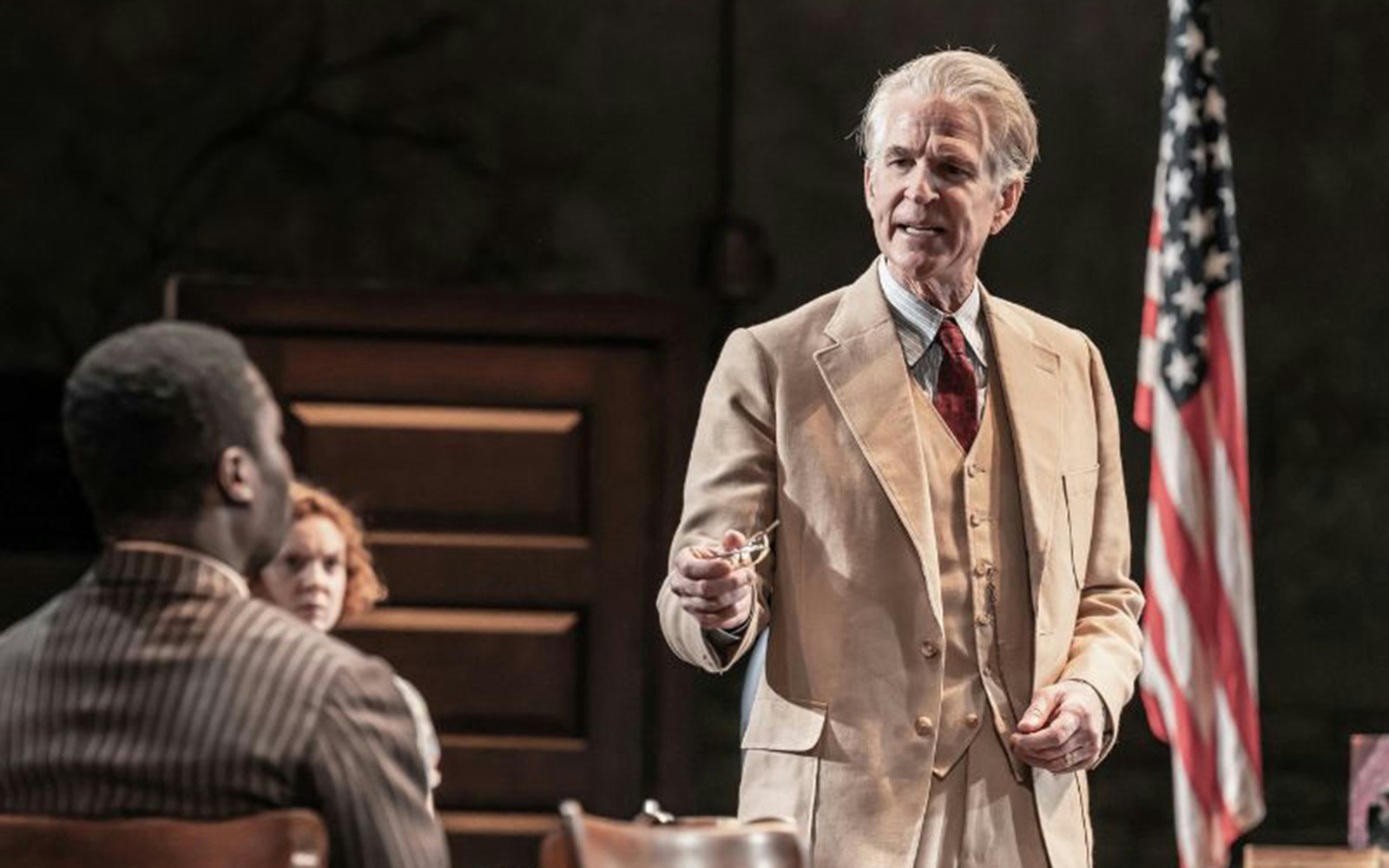 Courtroom scene from "To Kill a Mockingbird" play with a lawyer speaking, American flag in background.