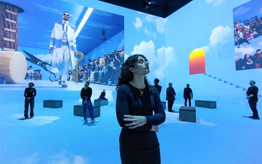Visitors at Vogue: Inventing the Runway exhibition, observing fashion projections in a blue-lit room.