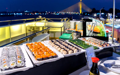 Buffet dinner with sushi on Chao Phraya cruise, Bangkok skyline in background.