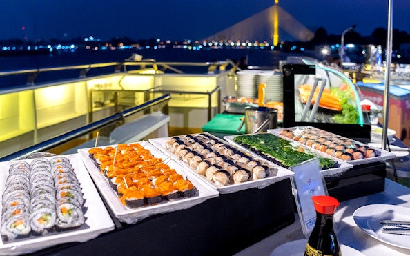 Buffet dinner with sushi on Chao Phraya cruise, Bangkok skyline in background.