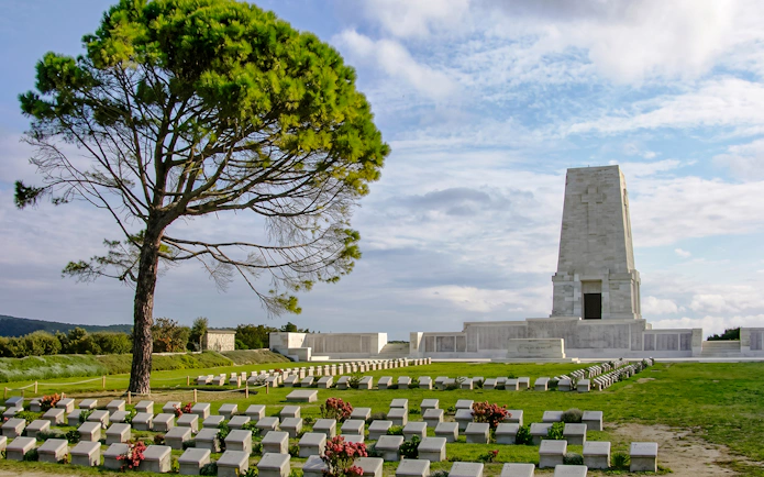 Gallipoli memorial site with gravestones and a lone tree under a cloudy sky.