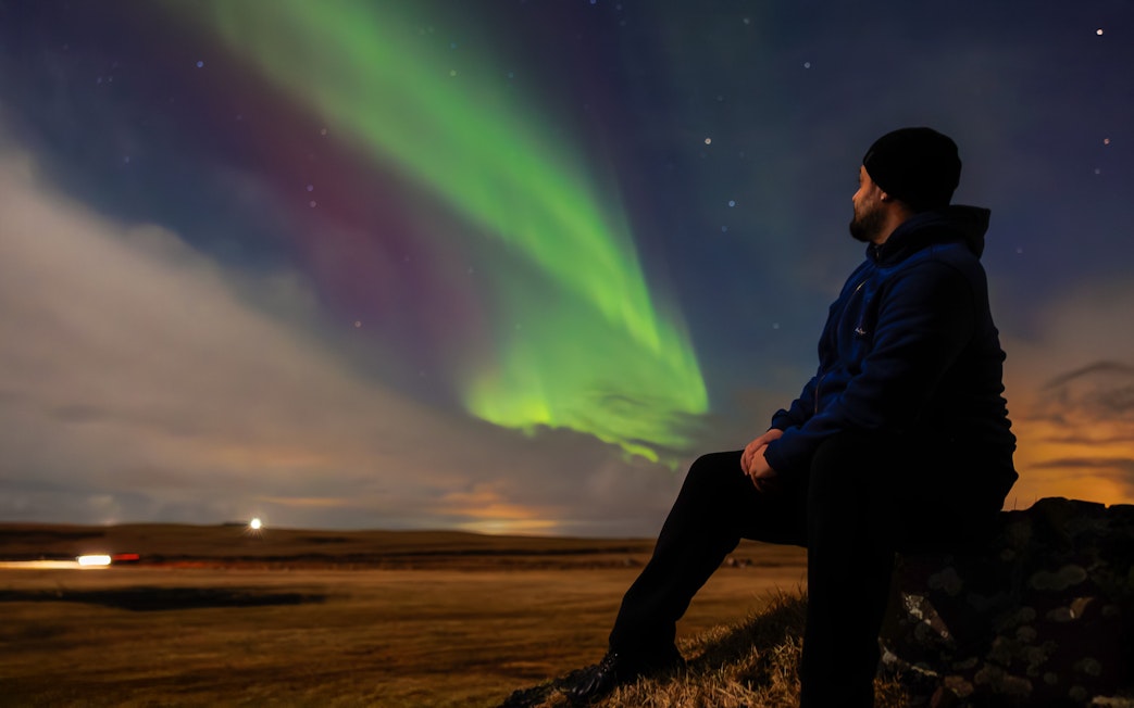 Person sitting on a rock watching green aurora borealis in the night sky.