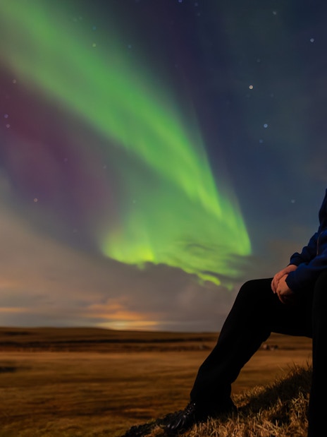 Person sitting on a rock watching green aurora borealis in the night sky.