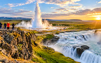 Geyser erupting near waterfalls in Iceland with tourists observing.