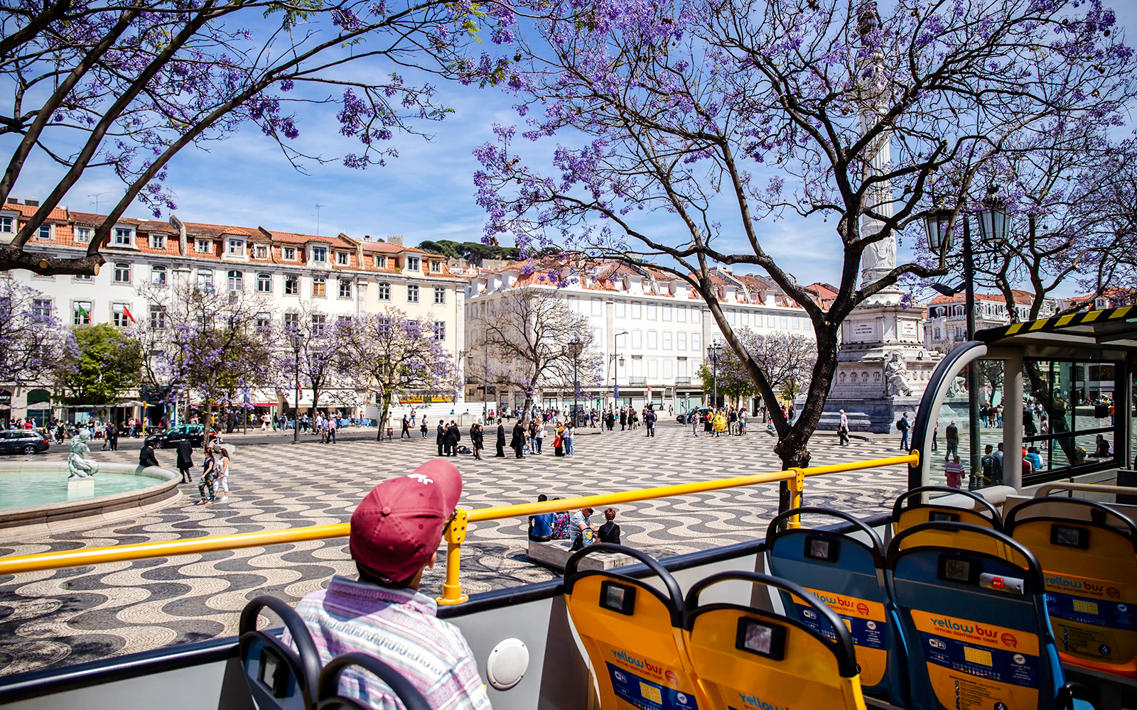 Tourists on yellow bus viewing Lisbon's Rossio Square with jacaranda trees.