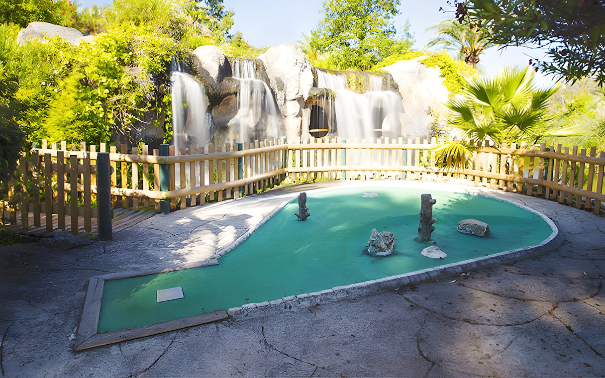 Mini-golf course with waterfall and wooden fence in a lush garden setting.