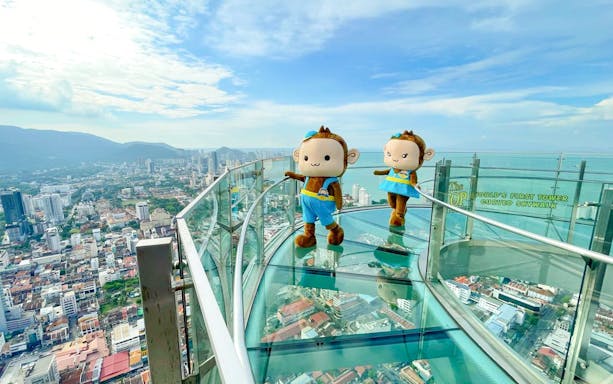 Skywalk view of Penang city with two monkey mascots on glass platform.