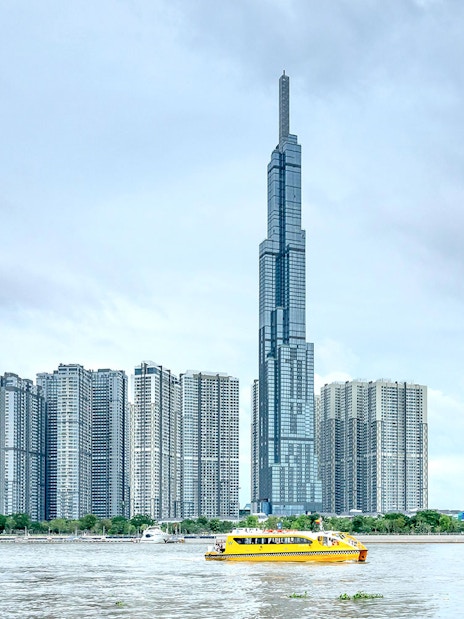 Landmark 81 and nearby skyscrapers by the river with a yellow ferry in Ho Chi Minh City.