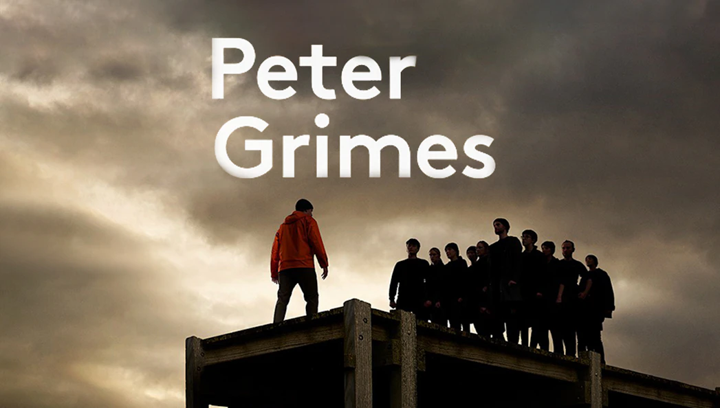 Man in orange jacket facing group on platform under cloudy sky, Peter Grimes.