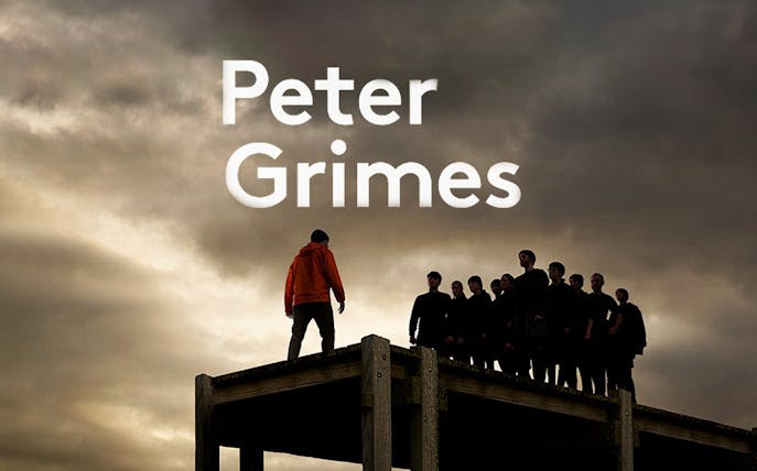Man in orange jacket facing group on platform under cloudy sky, Peter Grimes.