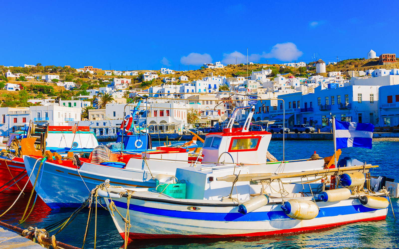 Fishing boats docked at the Old Port in Mykonos, Greece, with white buildings in the background.