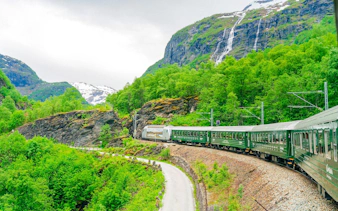 Flam railway train winding through lush green mountains with a waterfall in Norway.