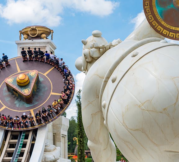 Visitors on a spinning ride at Parc Astérix, Paris, with a large statue in the foreground.
