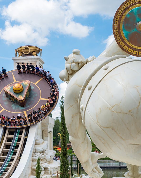 Visitors on a spinning ride at Parc Astérix, Paris, with a large statue in the foreground.