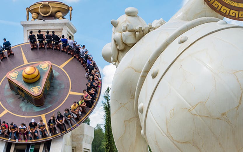 Visitors on a spinning ride at Parc Astérix, Paris, with a large statue in the foreground.