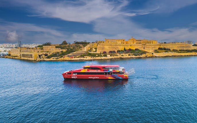 Red tour boat cruising near historic buildings in Valletta, Malta.
