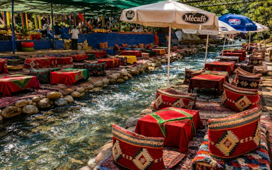 Outdoor dining setup by a stream in Ourika Valley, Marrakech, with colorful cushions and umbrellas.