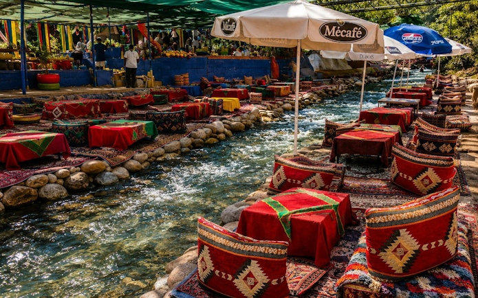 Outdoor dining setup by a stream in Ourika Valley, Marrakech, with colorful cushions and umbrellas.