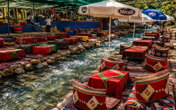 Outdoor dining setup by a stream in Ourika Valley, Marrakech, with colorful cushions and umbrellas.