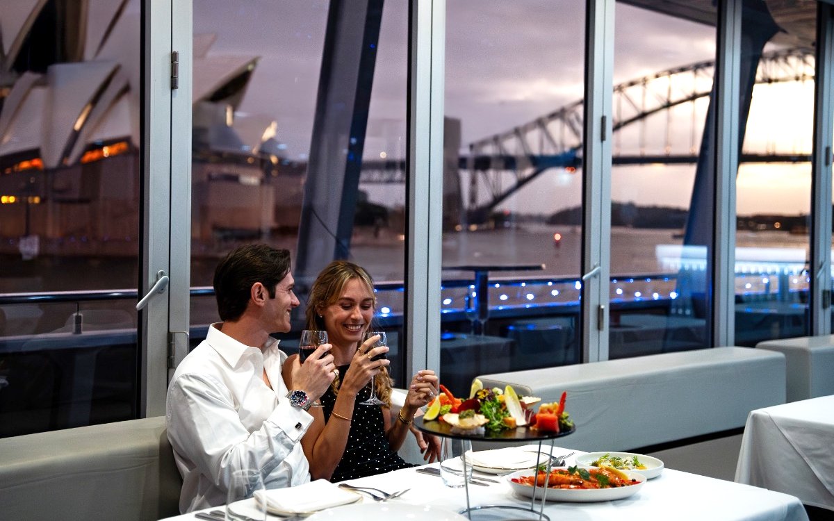 Couple enjoying dinner on a glass boat cruise with Sydney Opera House and Harbour Bridge views.