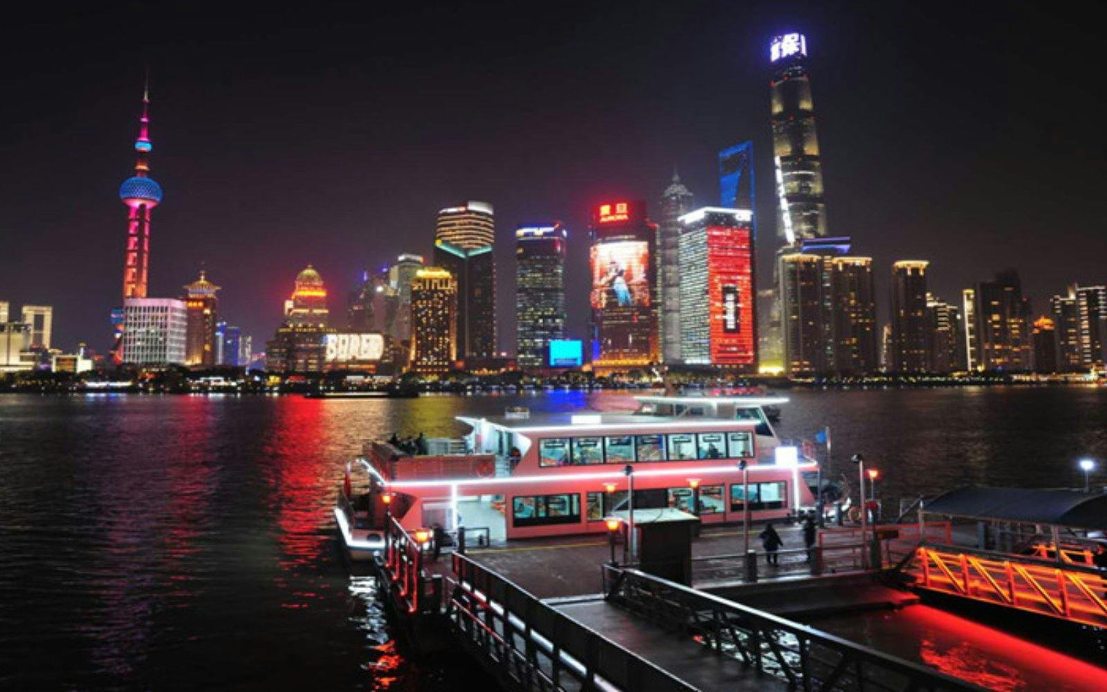 Cruise boat docked at Jinling East Road Pier with Shanghai skyline illuminated at night.