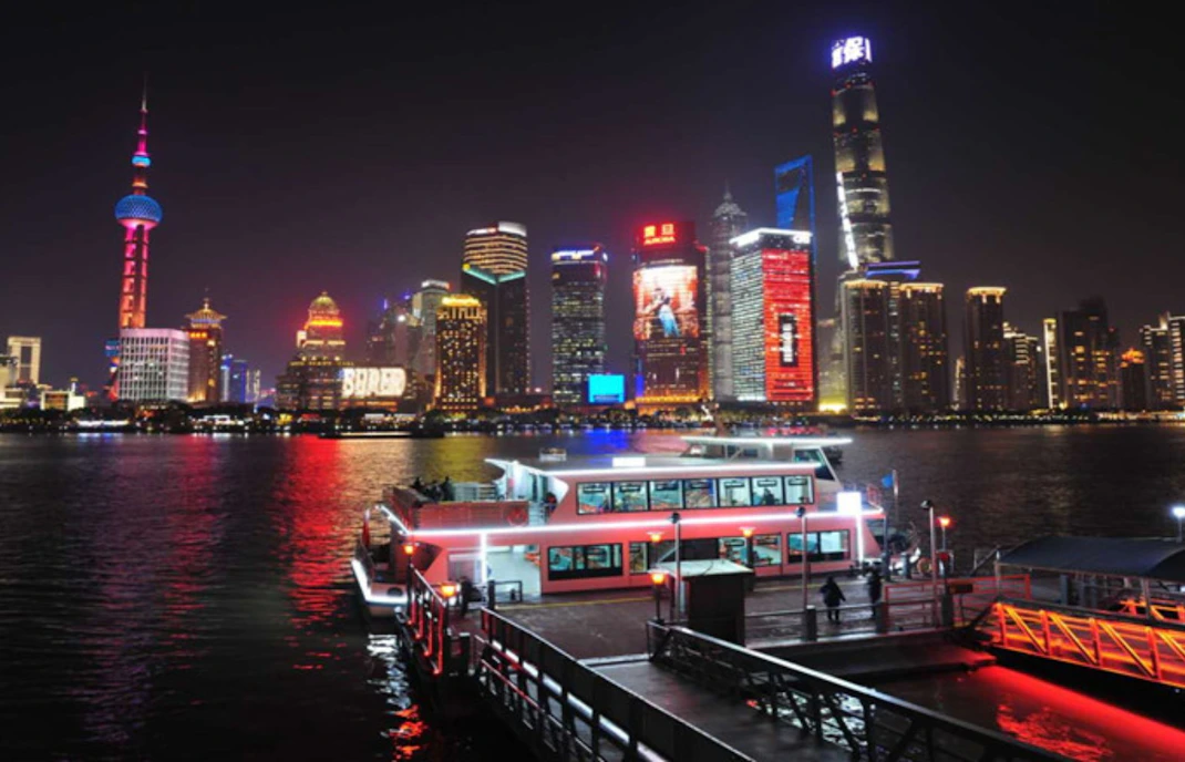 Cruise boat docked at Jinling East Road Pier with Shanghai skyline illuminated at night.
