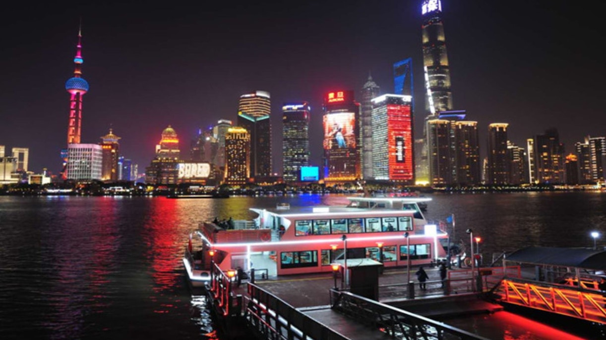 Cruise boat docked at Jinling East Road Pier with Shanghai skyline illuminated at night.