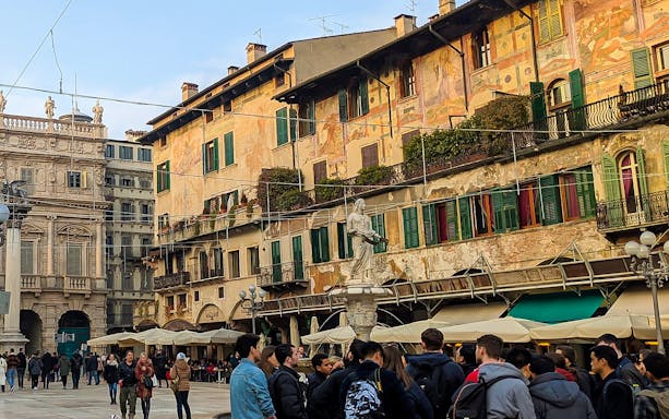 Tourists gather in Verona's Piazza delle Erbe, featuring historic buildings and a central statue.