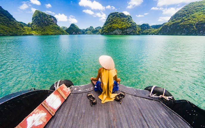 Person in traditional hat sitting on boat deck, Halong Bay limestone karsts in view.