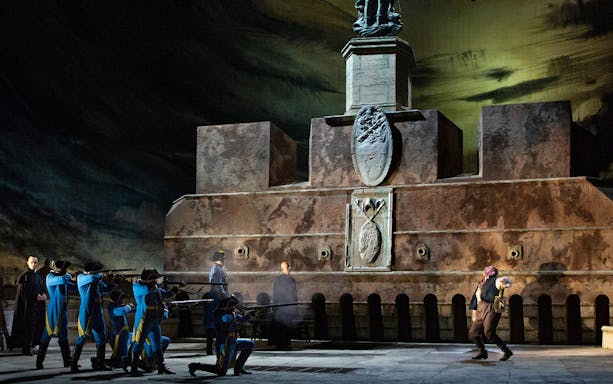 Opera scene from Tosca with soldiers aiming rifles at a man in front of a stone monument.