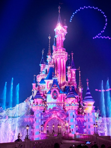 Disneyland Paris castle illuminated at night with colorful lights and fountain show.