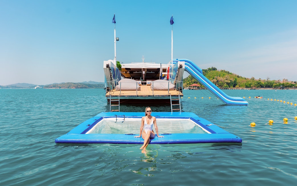 Inflatable floating pool with a person relaxing, yacht and slide in the background, tropical setting.