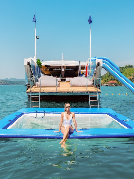 Inflatable floating pool with a person relaxing, yacht and slide in the background, tropical setting.