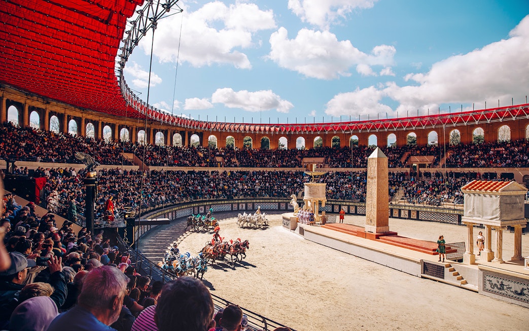 Chariot race in Puy du Fou arena with large audience and ancient Roman architecture.